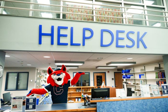 Blue, the college mascot, holding his arms out to highlight the library help desk sign, above the desk