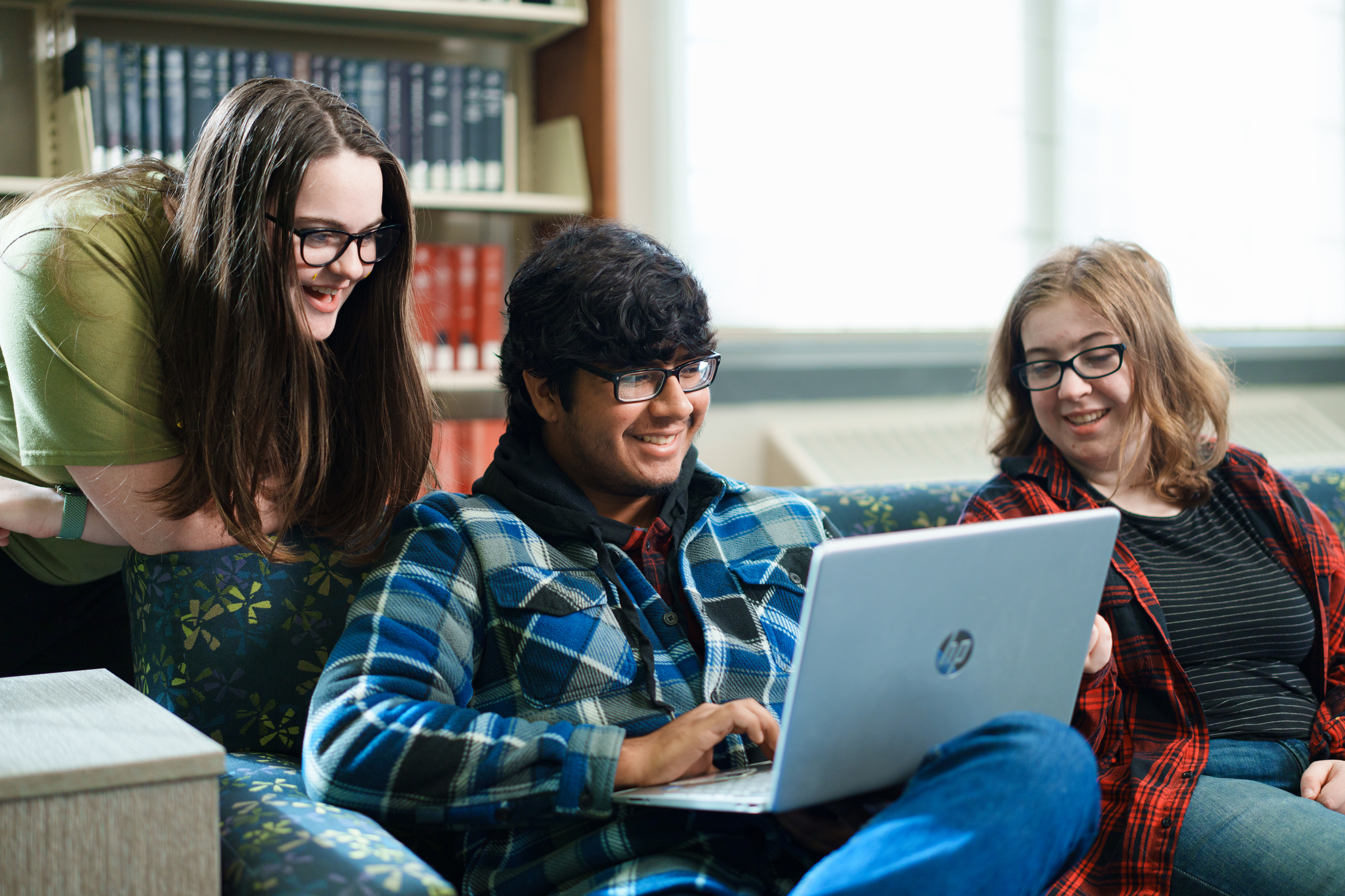 Two students on a library couch, one holding a laptop while the other looks on. Another student stands over their should laughing with them.